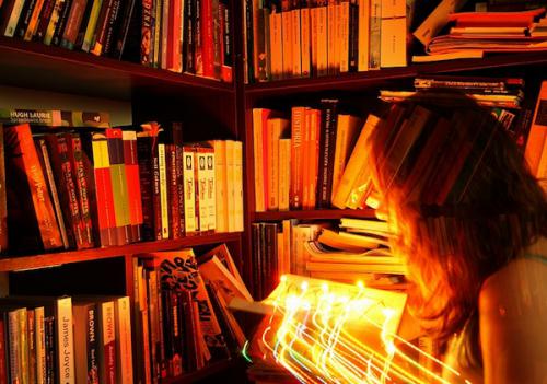 Standing among bookshelves, a woman holds an open book with bright lights shooting out of it