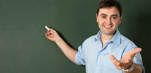 A teacher points to a chalkboard with chalk while reaching toward the viewer with other hand