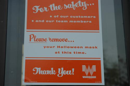 This photograph zooms in on a white and orange Whataburger sign that reads, "For the safety...of our customers and our team members. Please remove...your Halloween mask at this time. Thank You!"