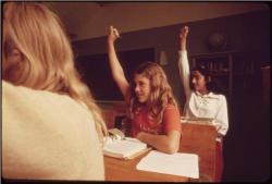 Three students sitting at desks with their hands raised